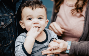 A baby with curly hair and striped shirt has fingers in mouth, held by an parent's hands.